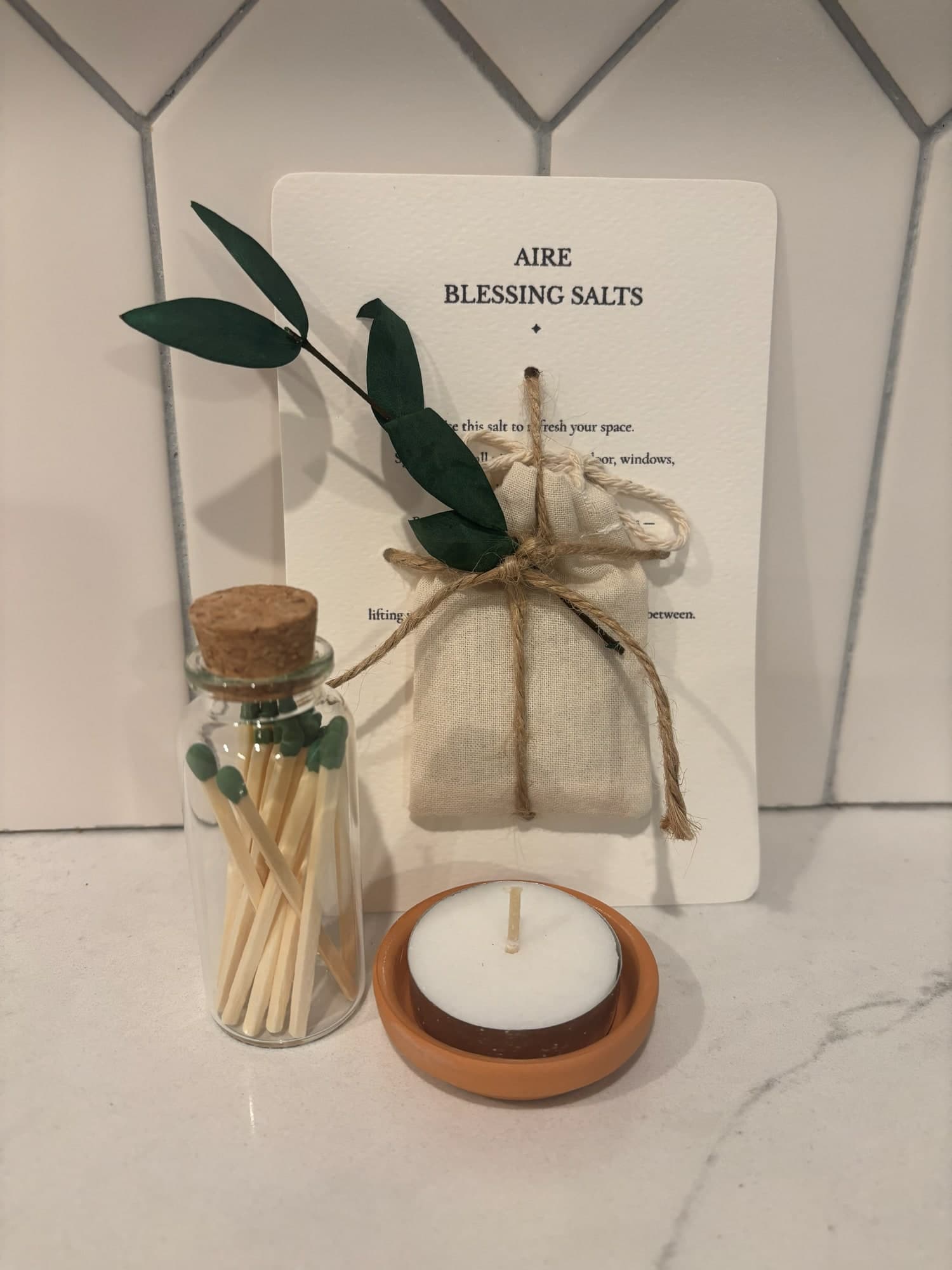 Aire blessing salts with a eucalyptus sprig on a printed card, a jar of matches and a tealight in a small terracotta dish