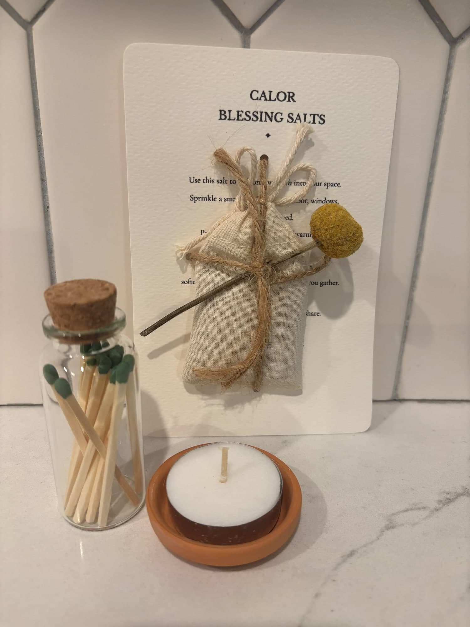 Calor blessing salts tied with twine and a yellow dried flower, beside a jar of matches and a tealight in a small terracotta dish