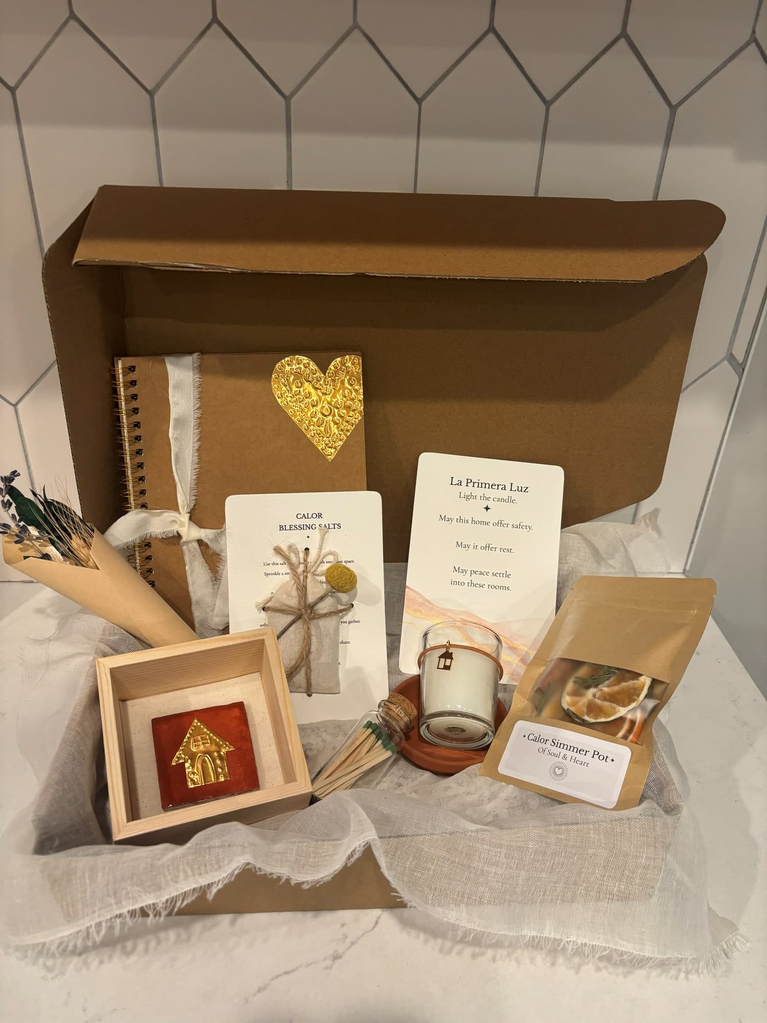 A Casa box packed for a new home, with a gold-heart journal, La Primera Luz card, Calor blessing salts, a framed terracotta house tile and a simmer pot